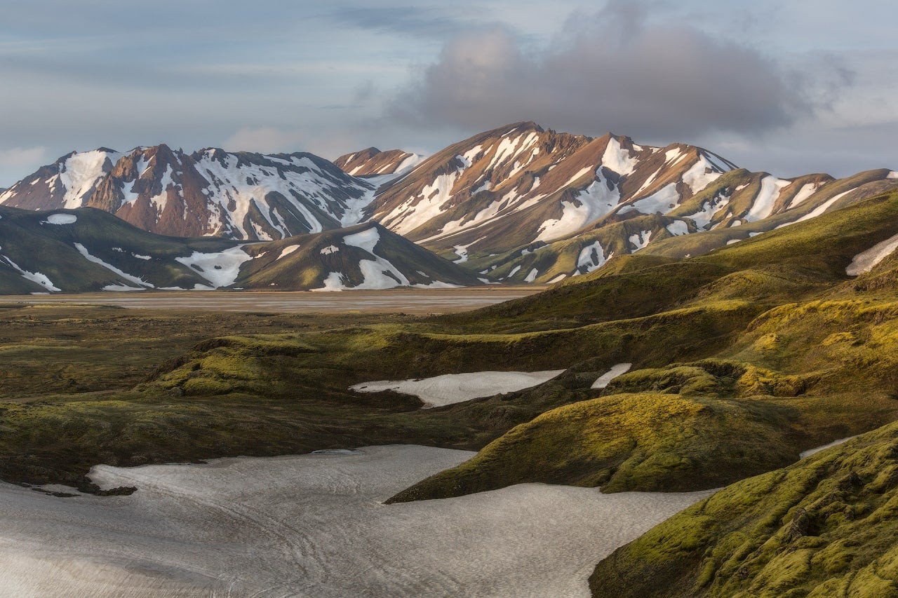 De fargerike toppene på Landmannalaugar lyser opp i et mykt lys, med grønn mose og smeltende snø i forgrunnen.