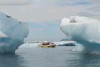 ヨークルスアゥルロゥン氷河湖（Jokulsarlon Glacier Lagoon）での水陸両用ボートツアー。ライフジャケットを着用した乗客が、2つの巨大な氷山の間に写っている。