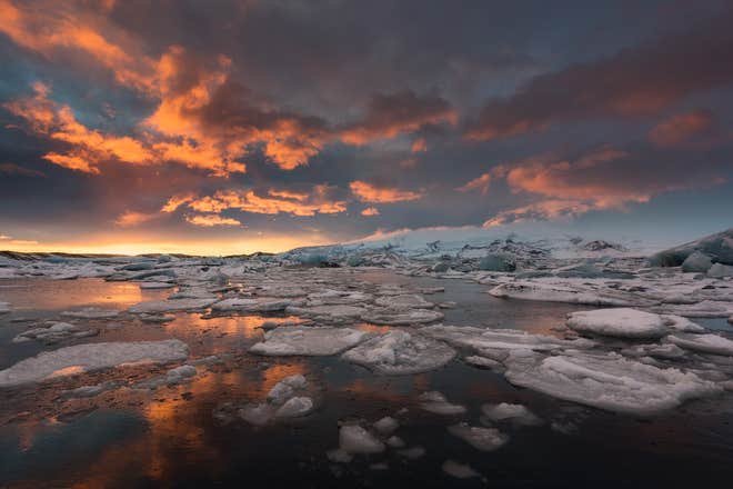 Sunset reflects off the icy waters of Jokulsarlon Glacier Lagoon, with floating icebergs under a dramatic sky.