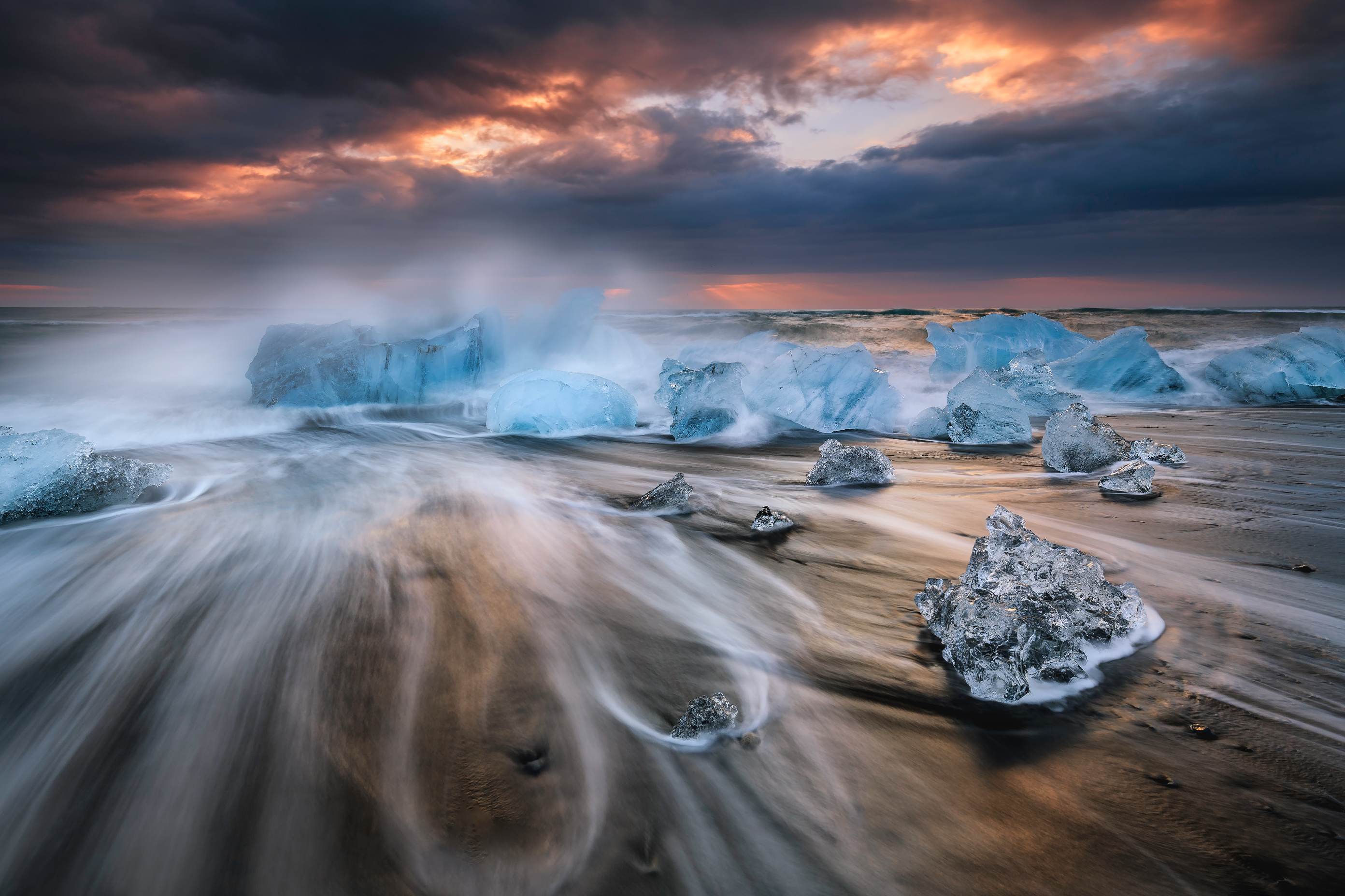 Las olas rodean trozos de hielo cristalino en la Playa de los Diamantes al amanecer, con nubes y una luz cálida brillando en el horizonte.