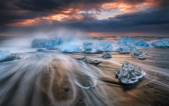Le onde si agitano intorno a pezzi di ghiaccio cristallino sulla Spiaggia dei diamanti all'alba, con le nuvole e la luce calda che brillano all'orizzonte.