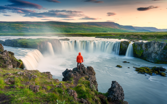 Un viaggiatore in giacca a vento davanti alla cascata di Godafoss al tramonto, durante il tour di 2 settimane in autonomia con auto a noleggio della Ring Road islandese.