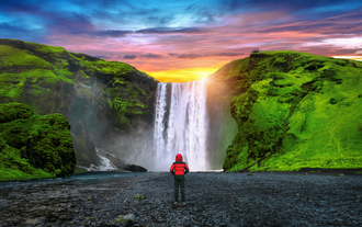 Viaggiatore con giacca rossa davanti alla cascata di Skogafoss, incorniciata da verdi scogliere e da un colorato cielo al tramonto nel sud dell'Islanda.