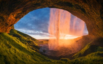 Der Wasserfall Seljalandsfoss leuchtet im Licht der untergehenden Sonne orange, wenn man ihn vom Höhlenpfad hinter den Wasserfällen aus betrachtet.