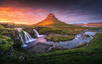 Sonnenuntergang über dem Berg Kirkjufell und dem Wasserfall Kirkjufellsfoss auf der Halbinsel Snaefellsnes mit leuchtendem Himmel und grüner Landschaft.