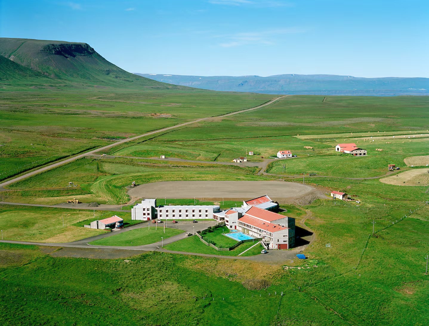 Aerial view of Hunavellir Guesthouse surrounded by open green fields, nearby cabins, and distant mountains under a clear sky.