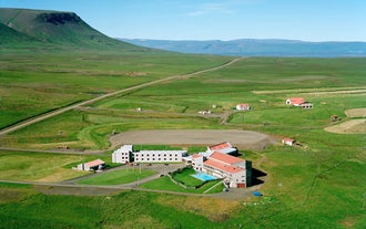 Aerial view of Hunavellir Guesthouse surrounded by open green fields, nearby cabins, and distant mountains under a clear sky.