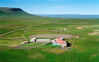 Aerial view of Hunavellir Guesthouse surrounded by open green fields, nearby cabins, and distant mountains under a clear sky.