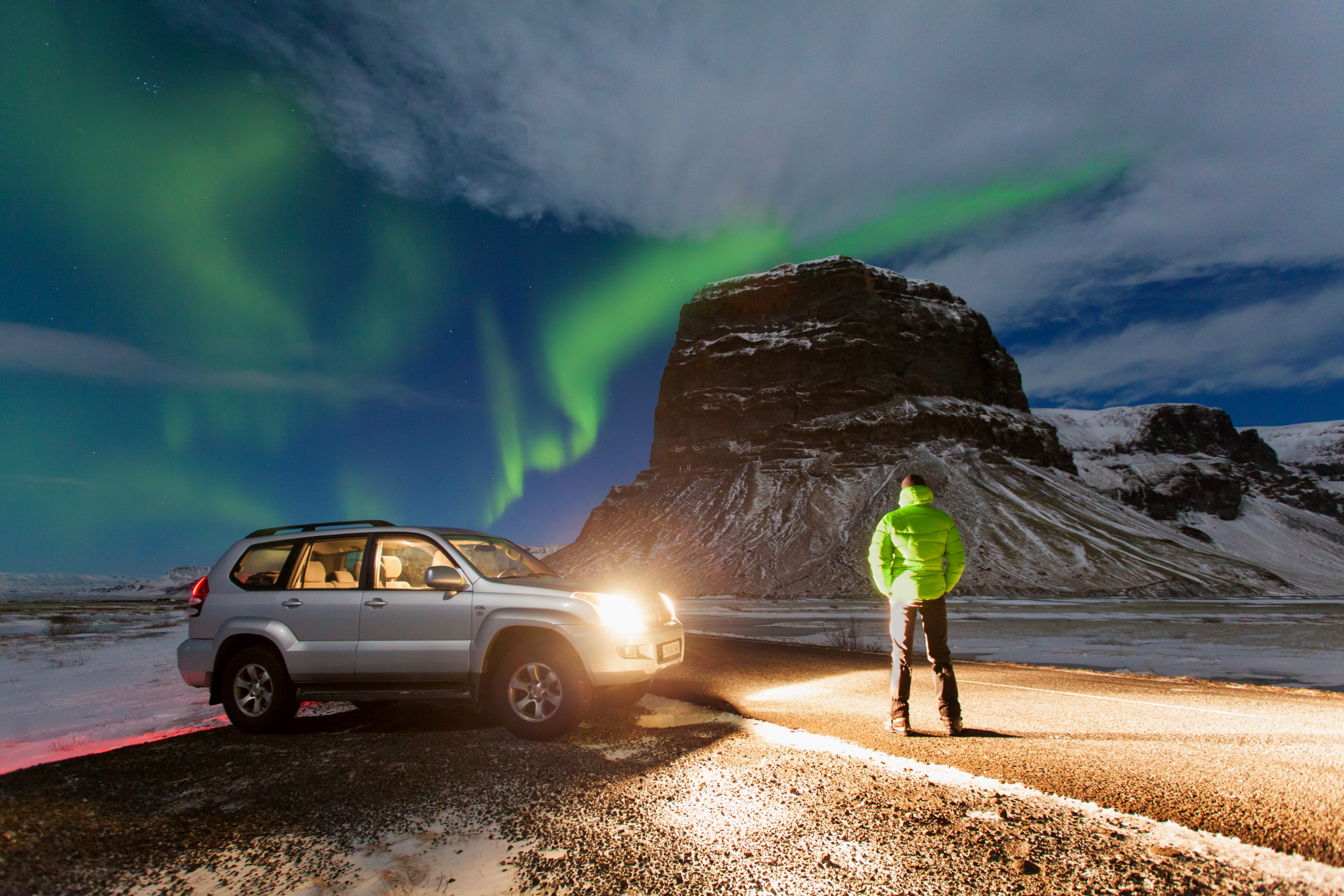A traveler watches the northern lights dance above Mount Lomagnupur in Iceland beside a car on a snowy roadside.