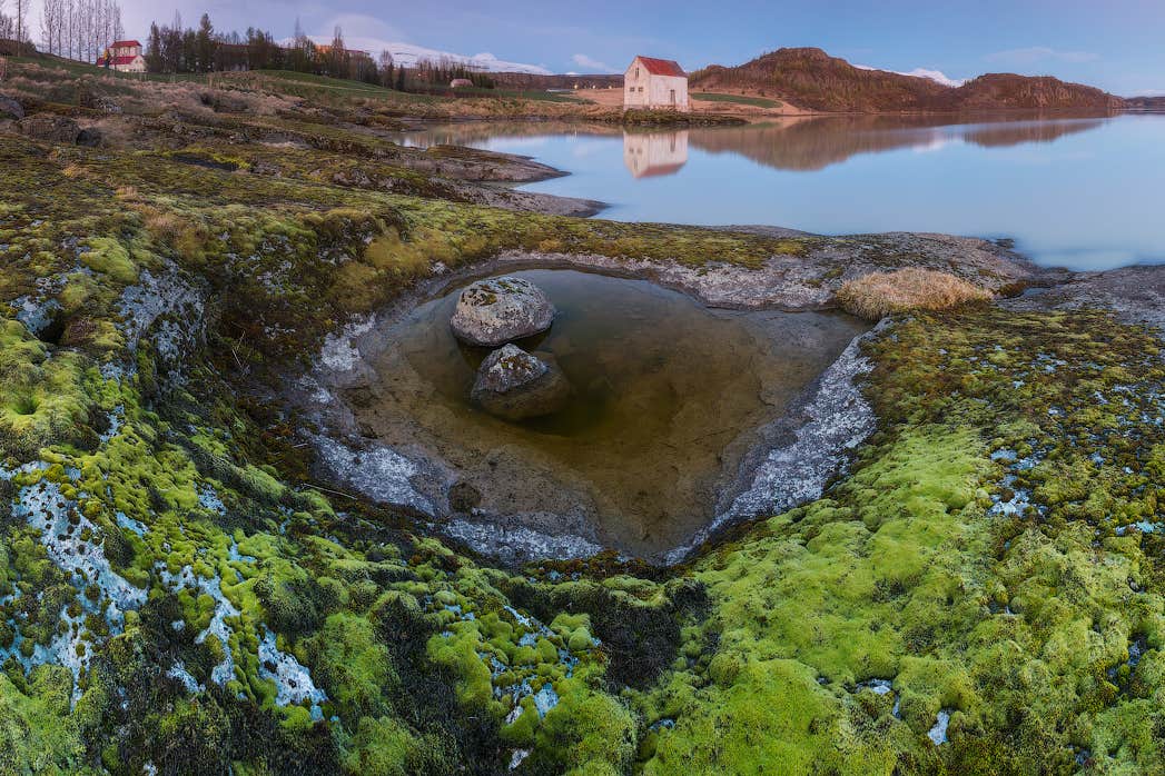 Un champ de lave couvert de mousse et un lac reflétant entourent une maison rustique près d’Egilsstadir, dans l’est de l’Islande, au coucher du soleil.