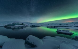 Noorderlicht gloeit boven het met sneeuw bedekte Jökulsárlón-gletsjermeer onder een met sterren gevulde hemel aan de zuidkust van IJsland.