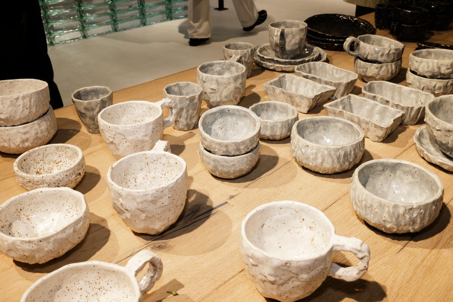 Display of handcrafted ceramic tableware in natural tones, featuring textured mugs, bowls, and plates arranged neatly on a wooden surface at a design store or market.