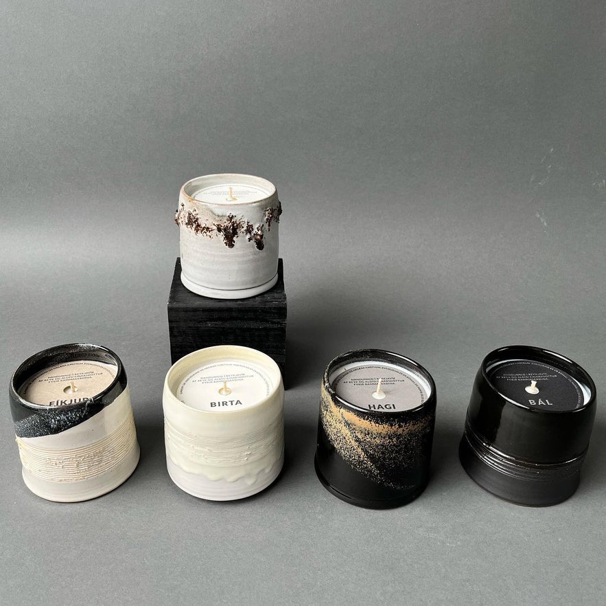 Assortment of Icelandic artisan candles in handcrafted ceramic jars, each with unique glazes and labeled scents including Eikjur, Birta, Hagi, and Bál, displayed against a gray background.