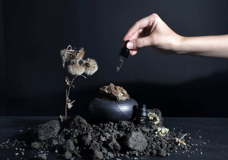 Hand using a dropper to apply Icelandic botanical serum from a black bottle labeled ILMOLIA, surrounded by volcanic rocks, dried flowers, and natural elements on a dark background.