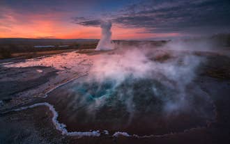 Le geyser Strokkur jaillit au coucher du soleil dans la vallée de Haukadalur en Islande, tandis que la vapeur s'élève d'un bassin géothermique voisin.