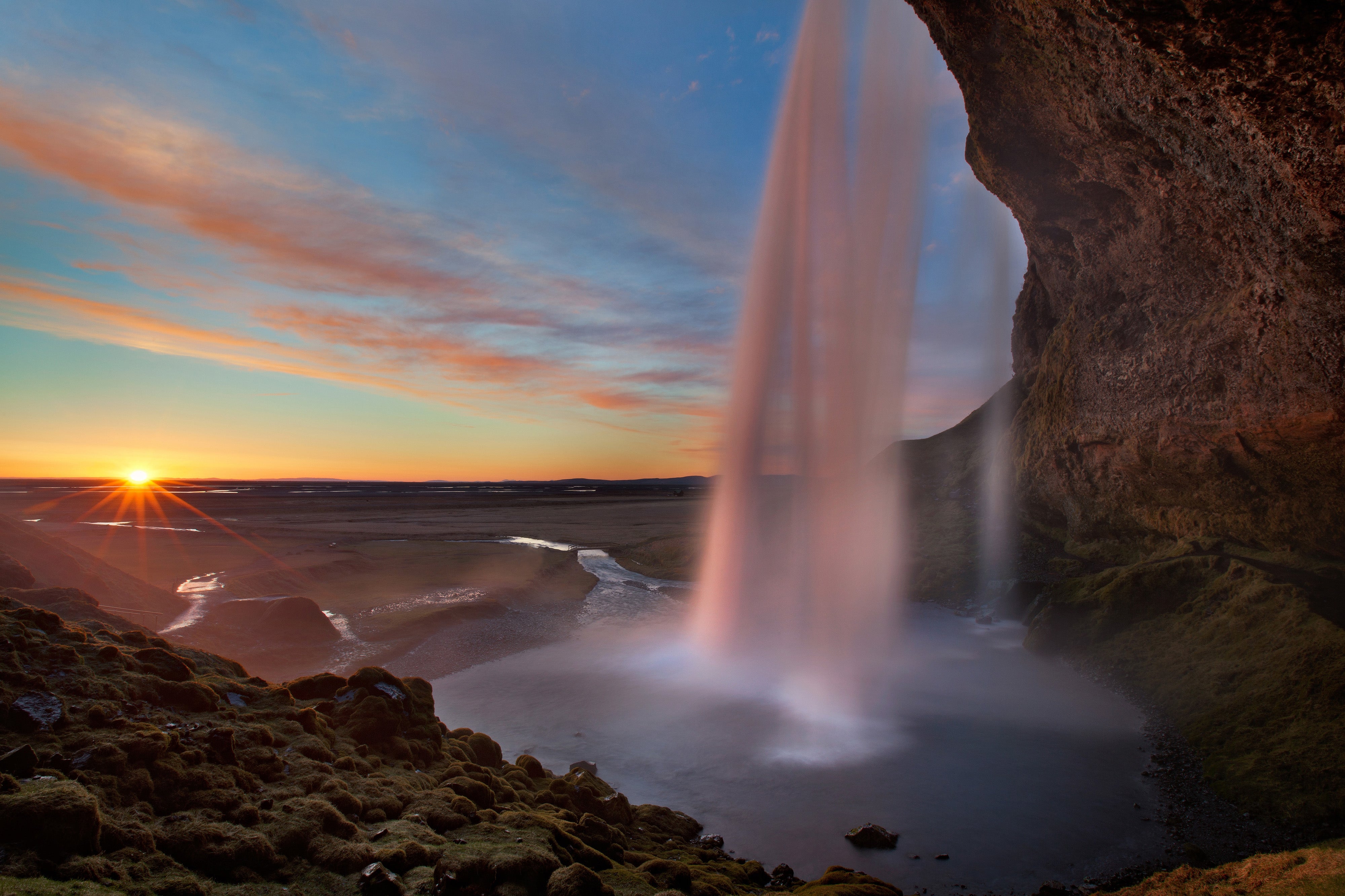 Seljalandsfoss waterfall at sunset, viewed from behind the cascade, with soft golden light and a winding river below.