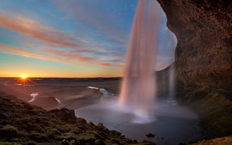 Seljalandsfoss waterfall at sunset, viewed from behind the cascade, with soft golden light and a winding river below.
