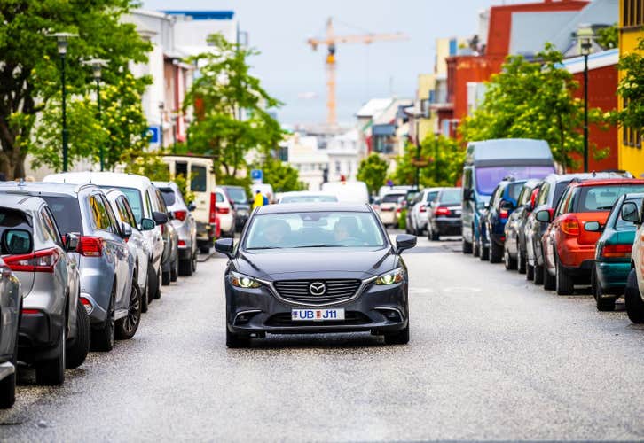 A private transfer car driving through a busy Reykjavik street lined with parked cars, trees, and colorful buildings in the background.