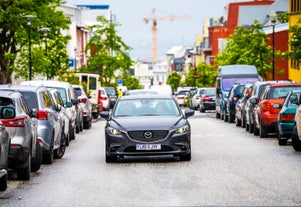 A private transfer car driving through a busy Reykjavik street lined with parked cars, trees, and colorful buildings in the background.