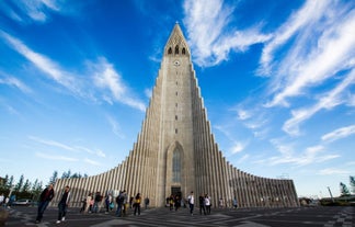 People walking in front of the tall tower of Hallgrimskirkja church on a cloudy blue-sky day.