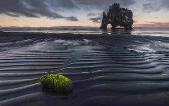 Moosbedeckter Felsen am schwarzen Sandstrand bei Ebbe mit dem Hvitserkur-Felsbogen in der Ferne bei Dämmerung.