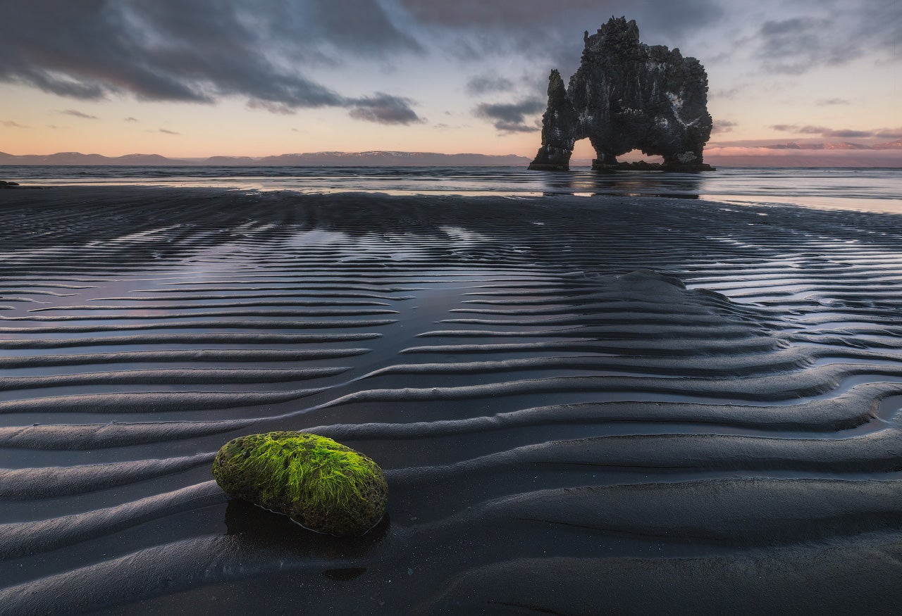 Roccia ricoperta di muschio su una spiaggia di sabbia nera con la bassa marea e il faro di Hvitserkur che si erge in lontananza al tramonto.