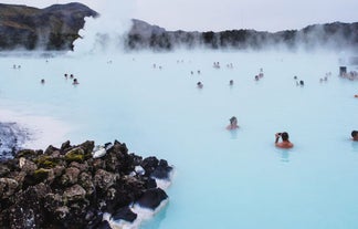People bathing in the milky-blue waters of the Blue Lagoon geothermal spa with a rocky natural landscape and steam rising into the air.
