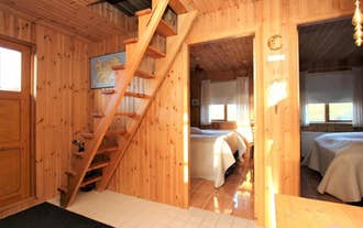 Wood-paneled cabin interior with steep wooden loft stairs, tiled flooring, and a cozy bedroom bathed in warm sunlight.