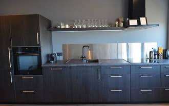 A compact kitchen with dark cabinets, a sink, a stovetop, and open shelves holding glasses and mugs.