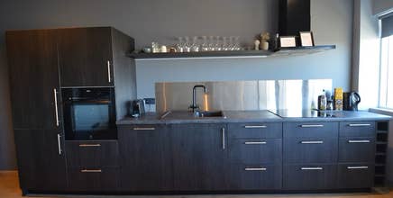 Cocina compacta con muebles oscuros, fregadero, placa de cocina y estantes abiertos con vasos y tazas.