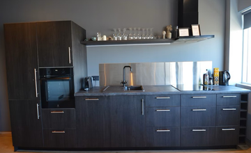 Et kompakt køkken med mørke skabe, en vask, en kogeplade og åbne hylder med glas og krus.