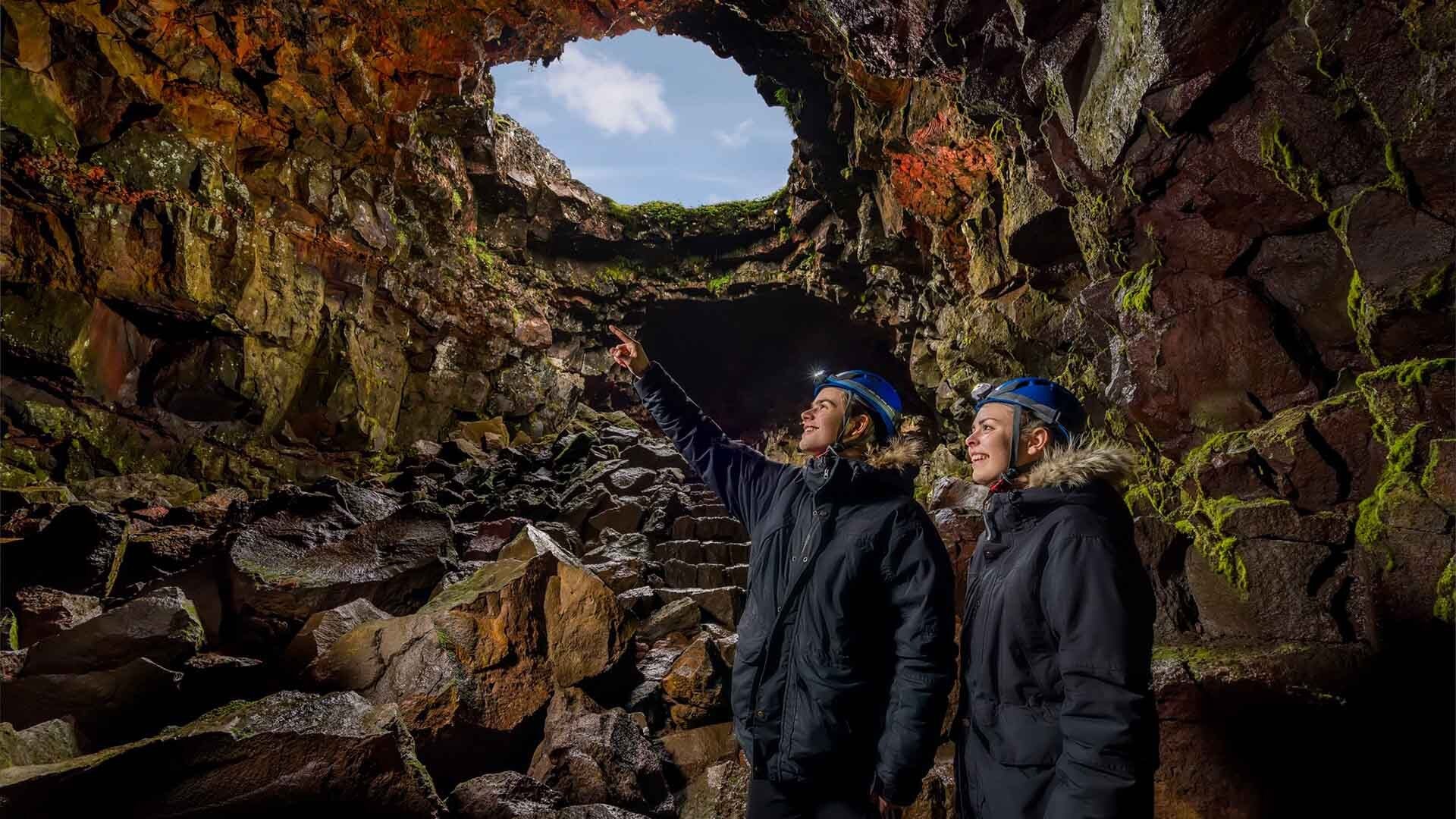 Two travelers wearing helmets explore Raufarholshellir Lava Tunnel, with one pointing toward lavacicles hanging from the jagged lava ceiling.