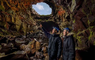 Two travelers wearing helmets explore Raufarholshellir Lava Tunnel, with one pointing toward lavacicles hanging from the jagged lava ceiling.