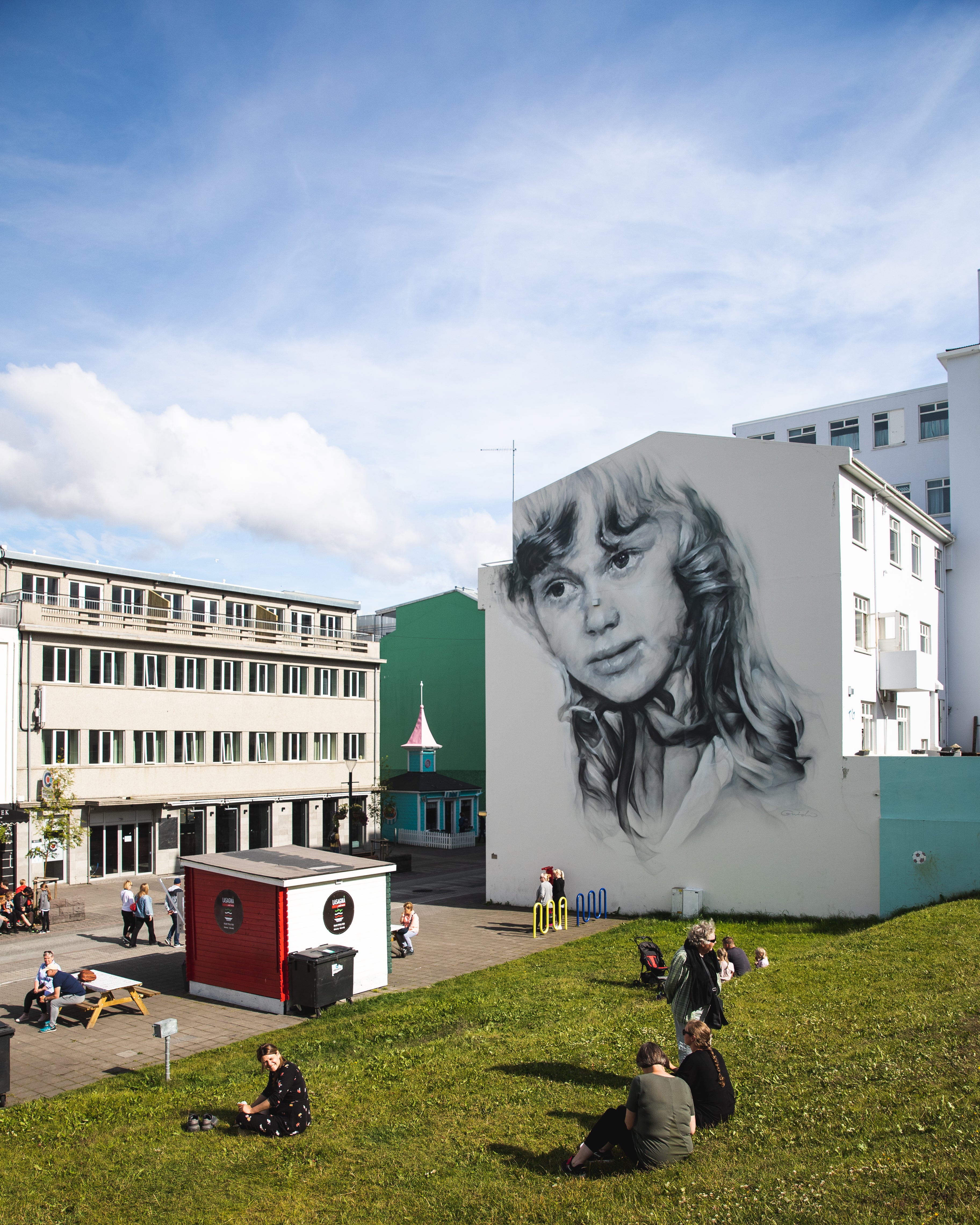 A large mural of a woman’s face decorates a building wall in Akureyri, with people relaxing in the square below.