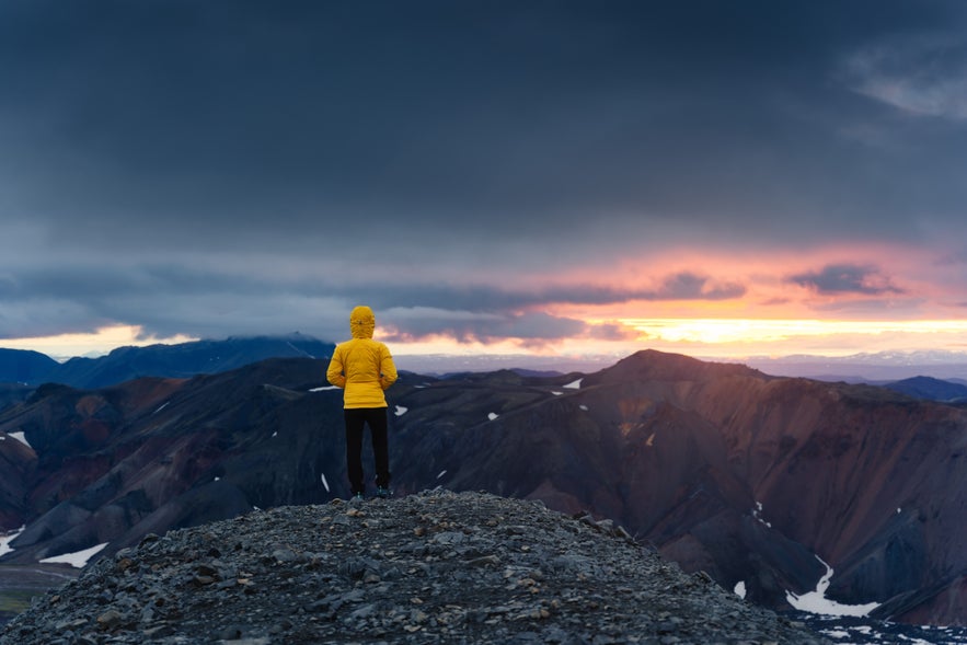 A woman hiker wearing a yellow jacket looking over the midnight sun in the Landmannalaugar-Hrafntinnusker hiking trail in Iceland’s Highlands.