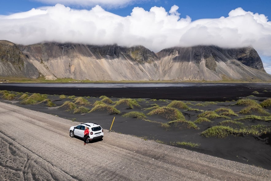 A car travels along a road in Stokksnes in Iceland amidst vast black sand plains framed by towering mountains Vestrahorn