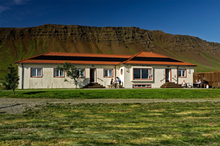 The exterior view of Reykholar HI Hostel with a grass area in front, the mountains directly behind, and people sitting around a 