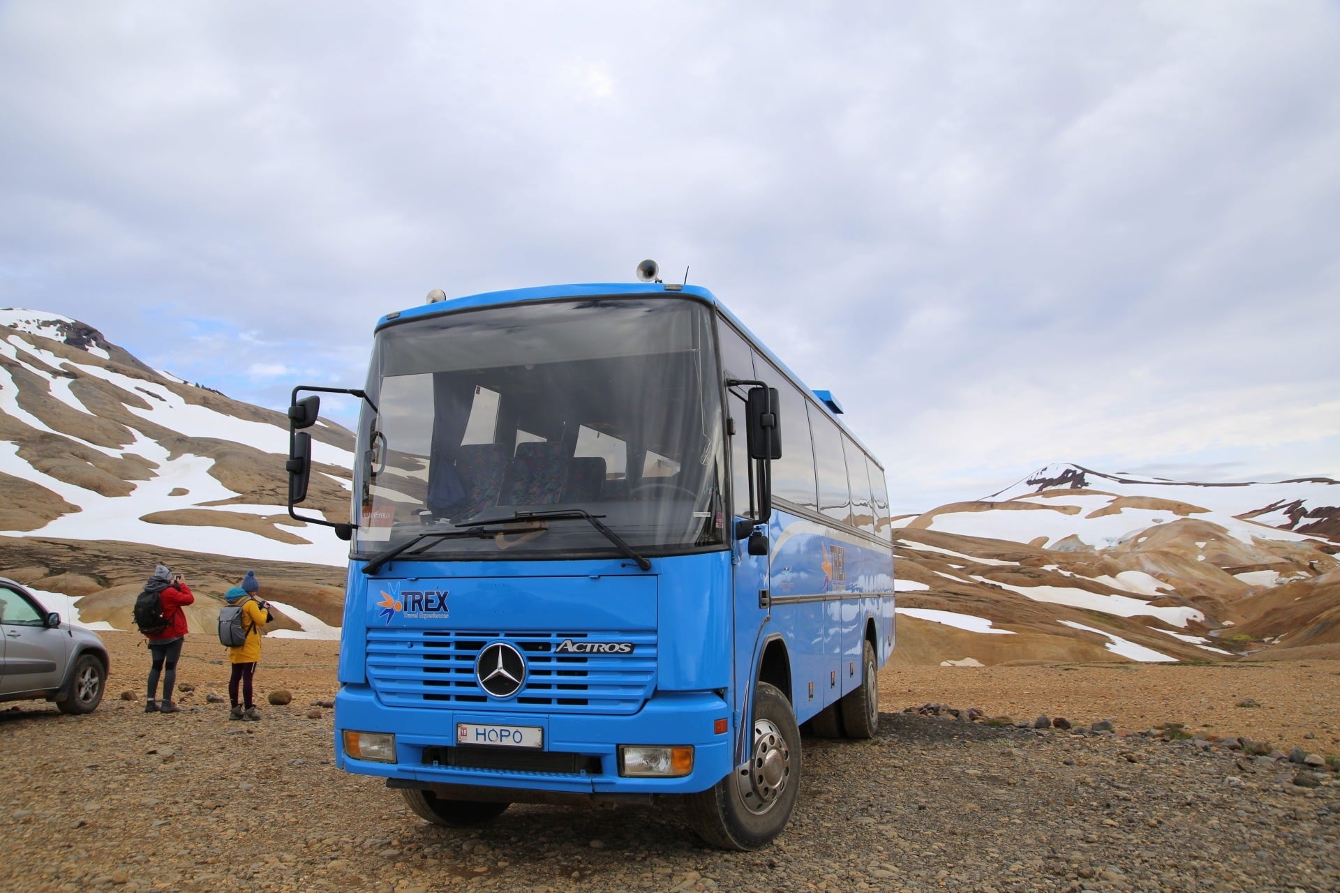 Blue highland bus parked in Iceland’s remote mountain region with snow patches, hikers nearby, and rugged terrain in the background.