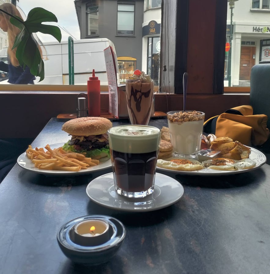 Brunch setup by the window at Prikið café with a burger, fries, coffee, yogurt, pancakes, and chocolate milkshake