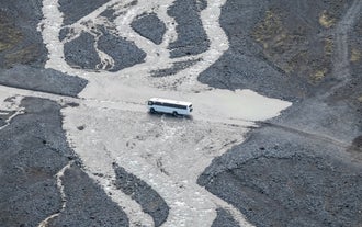 A bus crossing a shallow, rocky riverbed surrounded by black sand plains.