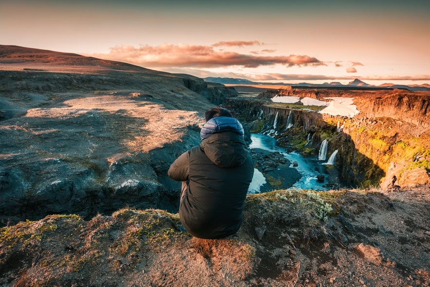 Rear view of traveler man sitting on edge of volcanic mountain with Sigoldugljufur waterfall on summer in Icelandic Highlands at evening