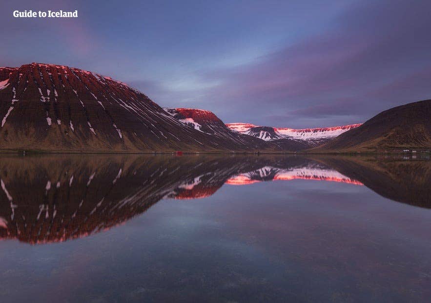 Sonnenuntergang über dem Isafjördur-Fjord in den Westfjorden Islands, mit schneebedeckten Bergen, die sich im ruhigen Wasser spiegeln.
