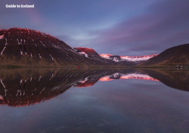 A beautiful view of Isafjordur with the mountains reflecting onto the deep blue water below, snow in the distance and a cloudy dark blue sky above.