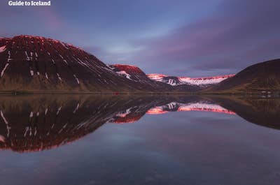 Une superbe vue sur Isafjordur avec les montagnes se reflétant dans l'eau bleu profond, la neige au loin et un ciel nuageux bleu foncé au-dessus.