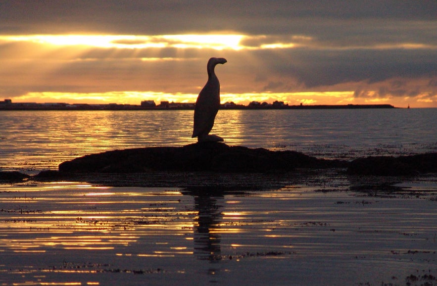 Silhouette of the Great Auk Memorial statue in Reykjavik, Iceland, standing on a coastal rock at sunset with golden light reflecting on the calm sea.