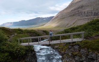 A hiker stands on a wooden bridge over a stream in the Westfjords of Iceland, with mountains and a rainbow in the background.