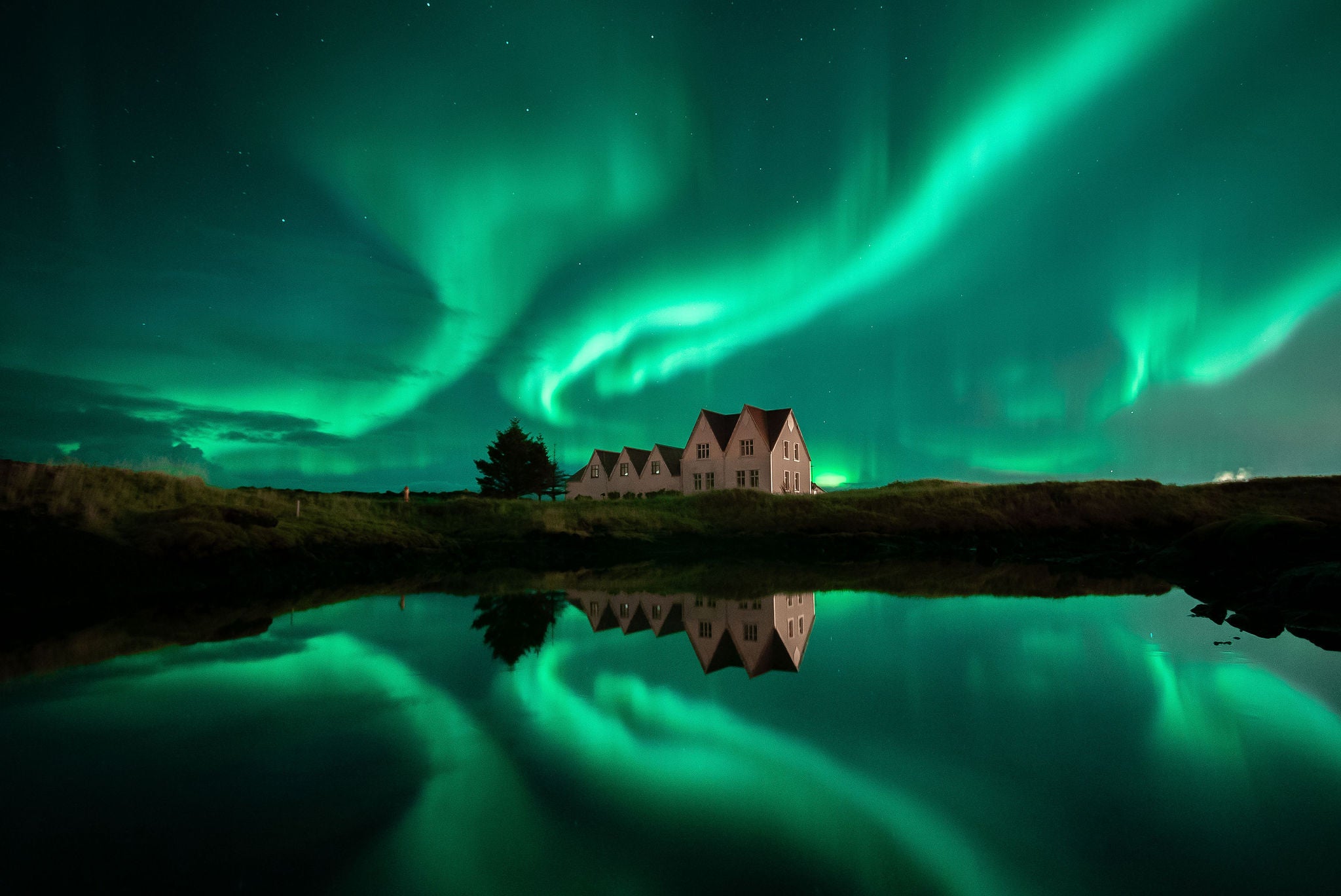 Northern lights glowing green above a countryside house in Iceland with reflections on a calm pond.