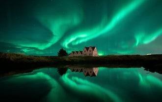 Northern lights glowing green above a countryside house in Iceland with reflections on a calm pond.