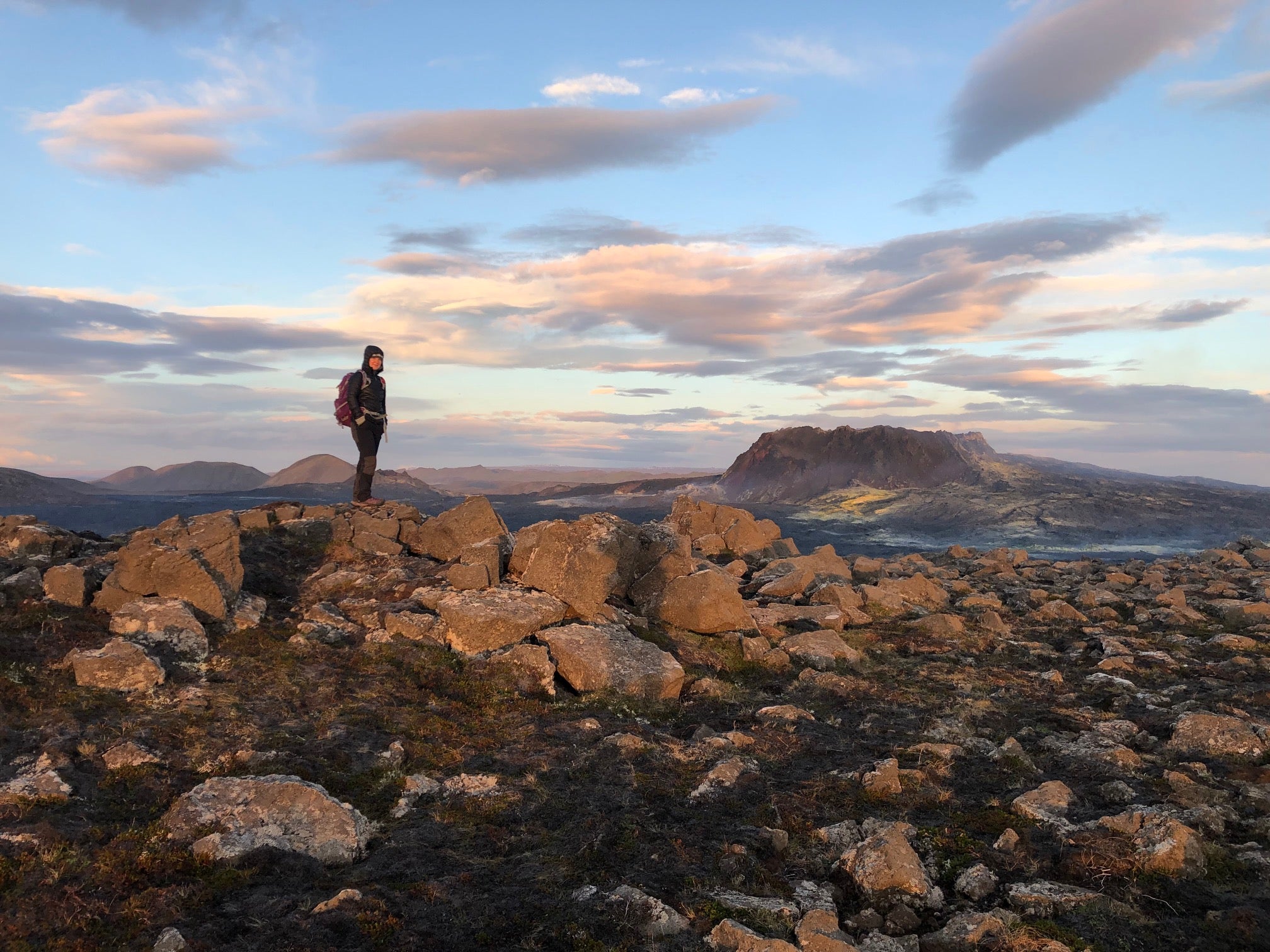 A hiker stands on rocky ground overlooking volcanic mountains on the Reykjanes Peninsula at sunset.
