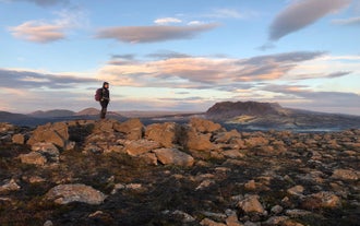 A hiker stands on rocky ground overlooking volcanic mountains on the Reykjanes Peninsula at sunset.
